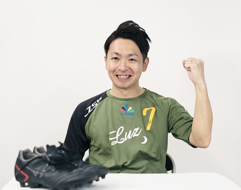 休日のT.M.さん。フットサルのユニフォーム姿で拳を上げ、机にスパイクを置いている