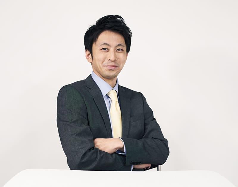 福祉用具営業のT.T.さん。スーツにネクタイ姿で腕を組んでいる